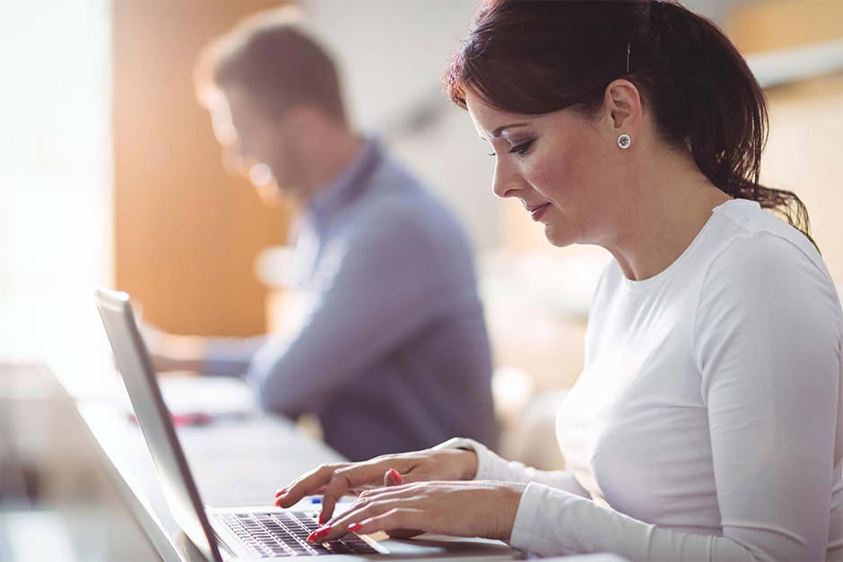 A woman is typing on her laptop while another man watches.